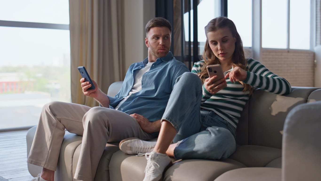 Offended couple browsing smartphones at domestic weekend. Sad boyfriend looking focused girlfriend hold mobile phone at couch. Digitally obsessed pair sitting sofa together ignoring each other at home